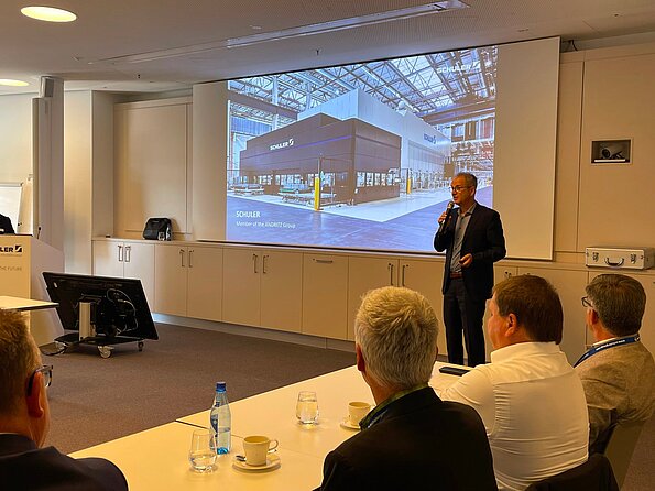 Präsentation in einem Konferenzraum: Ein Referent spricht mit Mikrofon vor einer Leinwand mit SCHULER-Produktionsanlage, mehrere Zuhörer sitzen am Tisch mit Getränken und Notizen im Vordergrund.