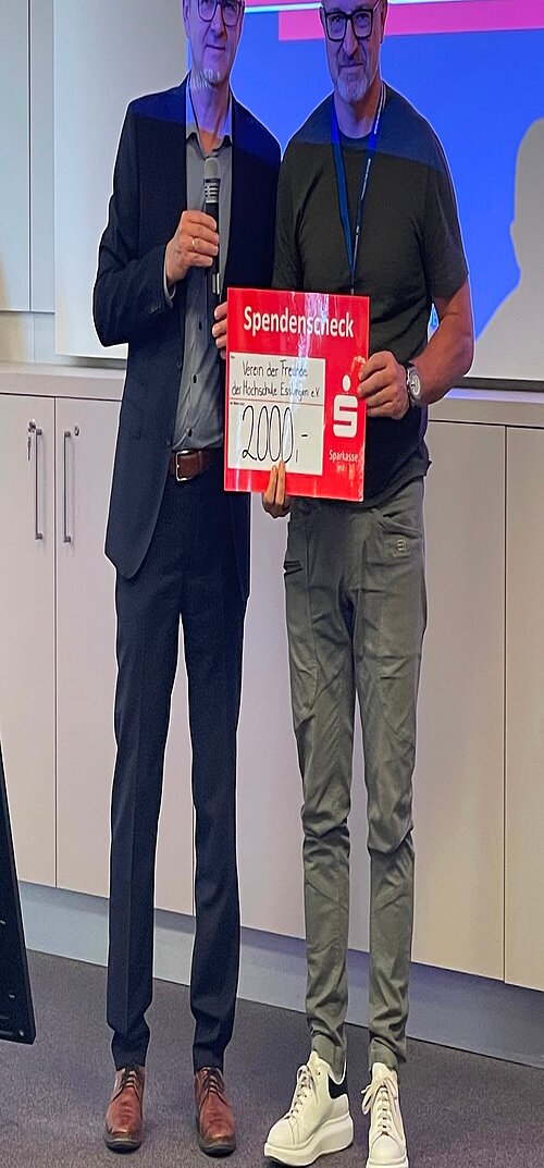 Zwei Personen bei einer Spendenübergabe mit symbolischem Scheck über 2.000 Euro vor einer Präsentationsfolie.