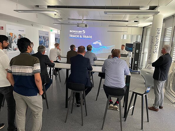 Mehrere Gäste verfolgen in einem modernen Schulungsraum die Präsentation „Track & Trace – Parts Tracking from Start to Finish“ von Schuler, während der Referent vorne vor der projizierten Folie steht.