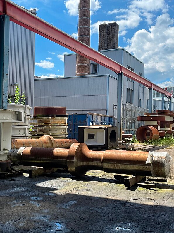 Große geschmiedete Industriewelle auf einem Werksgelände vor Produktionsgebäuden und Schornsteinen.