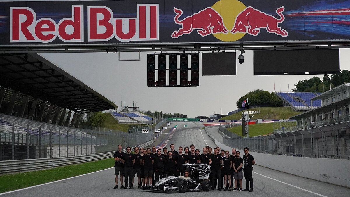 Gruppenfoto des Rennstall-Teams der Hochschule Esslingen, vor einer Rennstrecke. Im Vordergrund steht der Formelwagen.