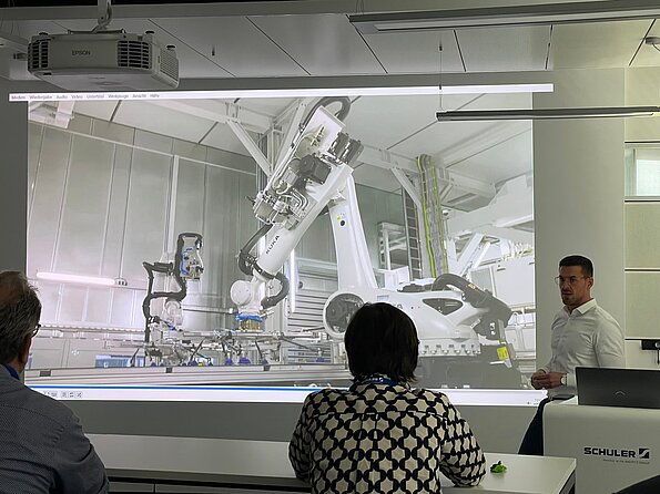 Referent präsentiert ein Video mit Industrierobotern in einer Fertigungsanlage, während mehrere Zuhörer im Seminarraum die Projektion verfolgen.