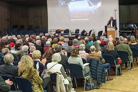 Gefüllter Saal mit applaudierenden Menschen, beim Matineekonzert des VdF Alumni + Friends, am Rednerpult wird das Konzert durch ein paar Begleitworte eingeleitet.