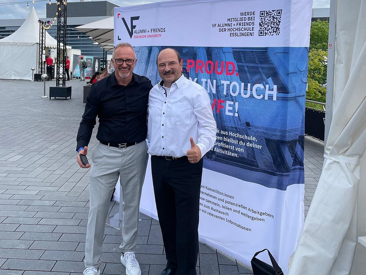 Zwei Personen posieren vor einem Roll-up-Banner von VDF Alumni + Friends bei einer Open-Air-Veranstaltung auf dem Gelände des Mercedes-Benz Museums.