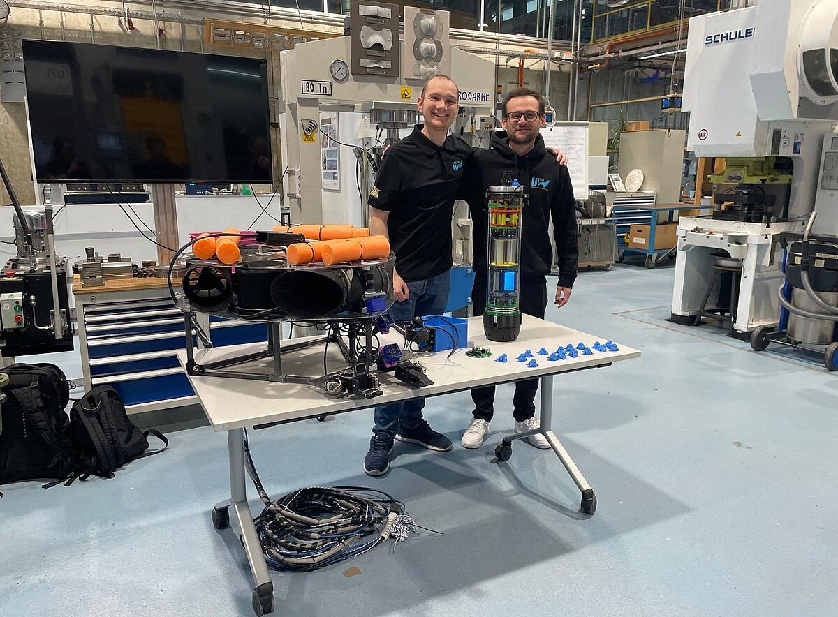 Zwei junge Männer stehen in einer Werkhalle neben dem technischen Prototypen einer semi-autonomen Tauchboje, auf einem Tisch.